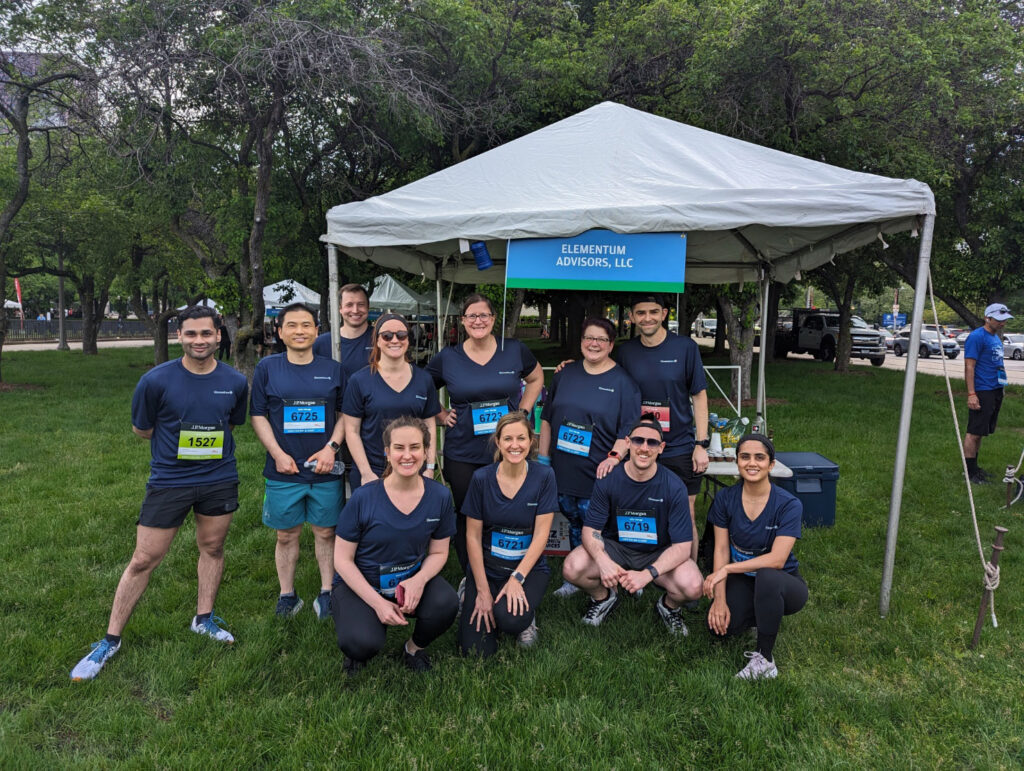 A group of twelve people in matching blue shirts pose in front of a tent labeled "Elementum Advisors, LLC" at an outdoor event, with trees and grass in the background.