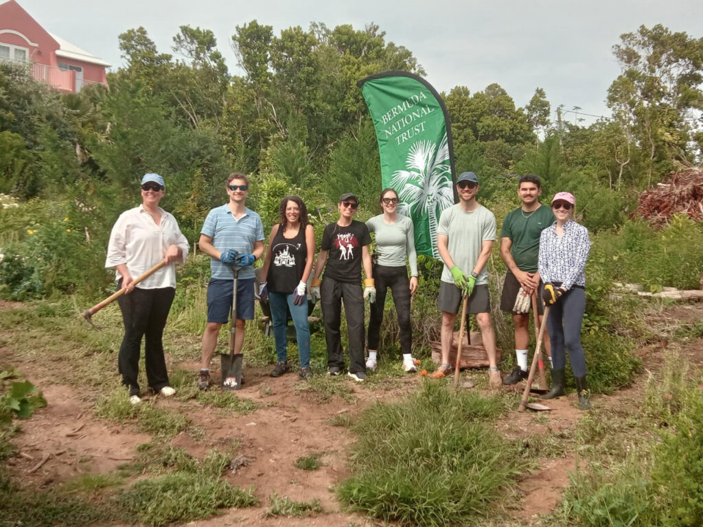 Elementum Advisors employees stand outdoors on grassy ground holding gardening tools, posing in front of a green "Bermuda National Trust" banner.