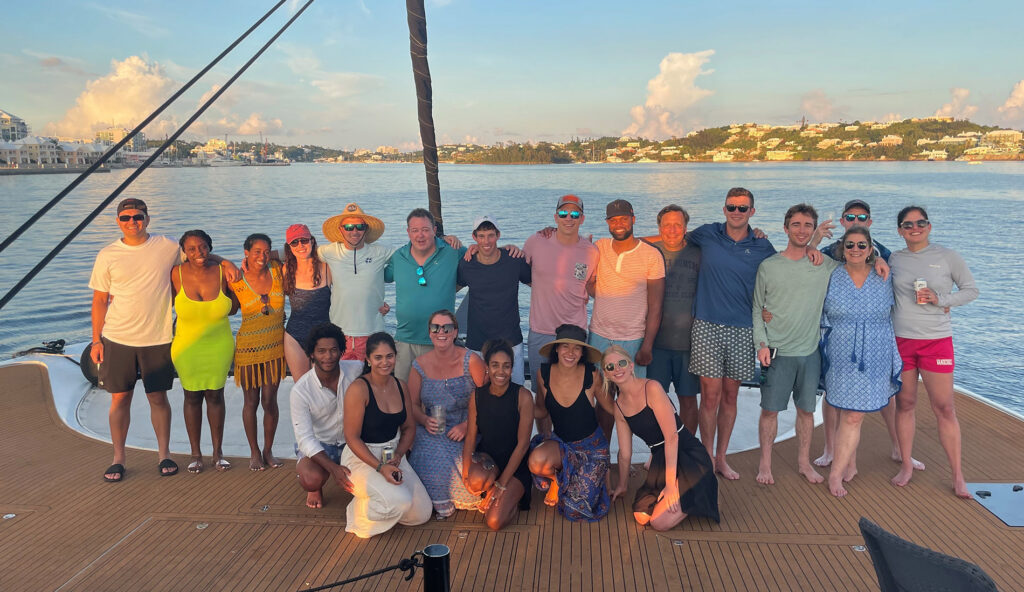 Elementum Advisors employees kneel on the deck of a boat, smiling at the camera with water and shoreline in the background