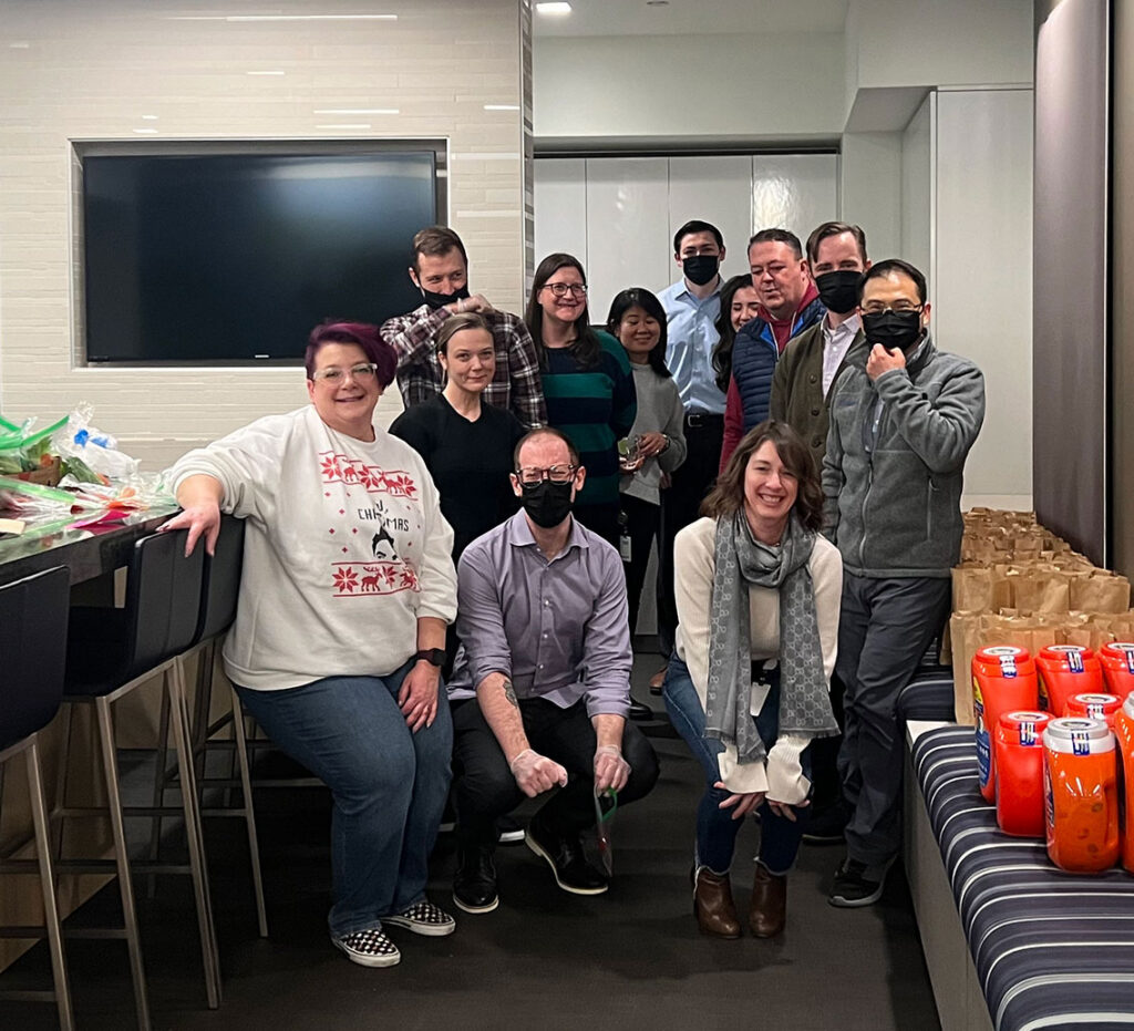 Elementum employees pose for a photo indoors near a counter with food items, brown paper bags, and several orange containers on the floor.